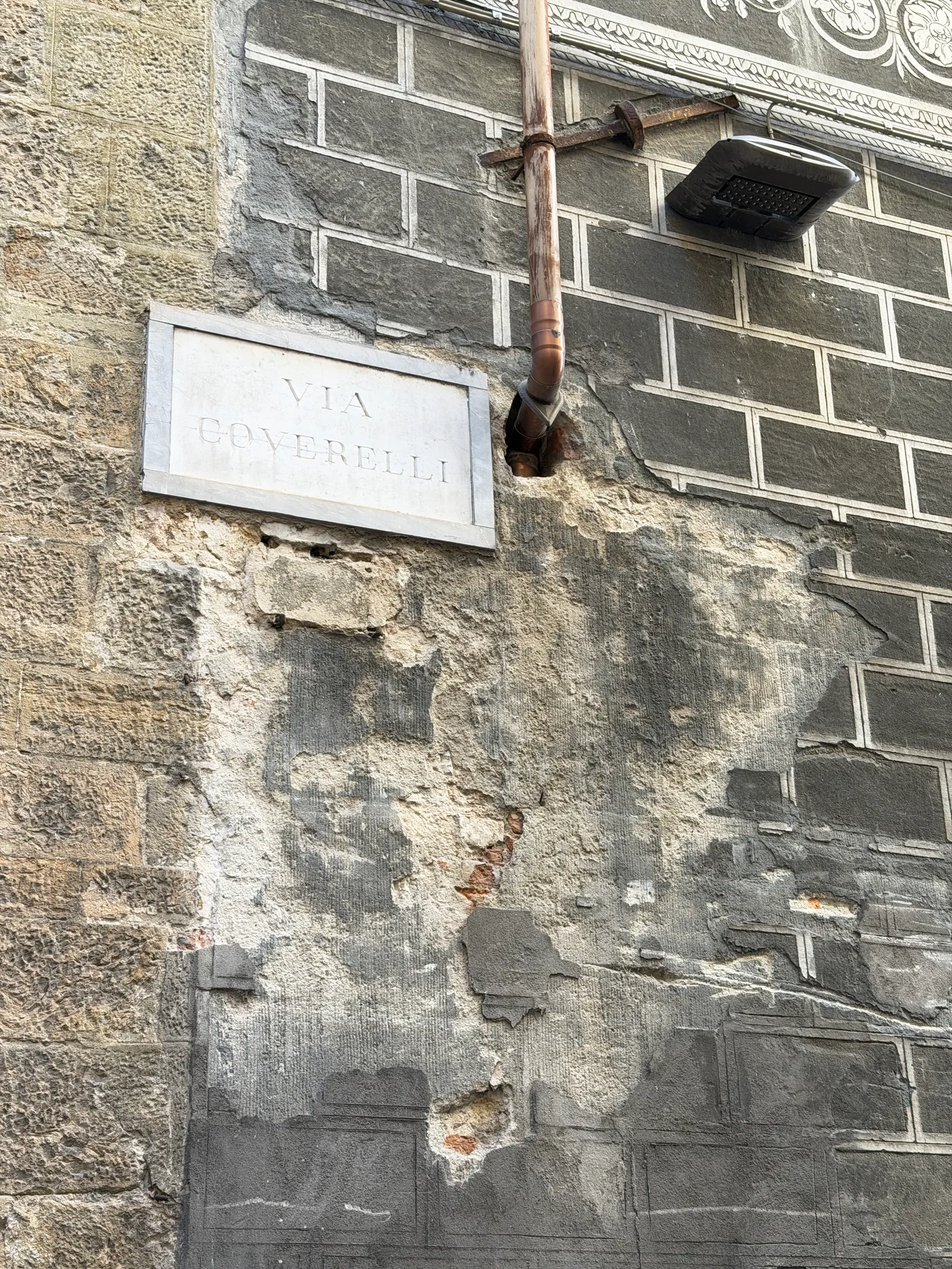 A marble street name plate set into a rough stone wall, the render broken away around it.