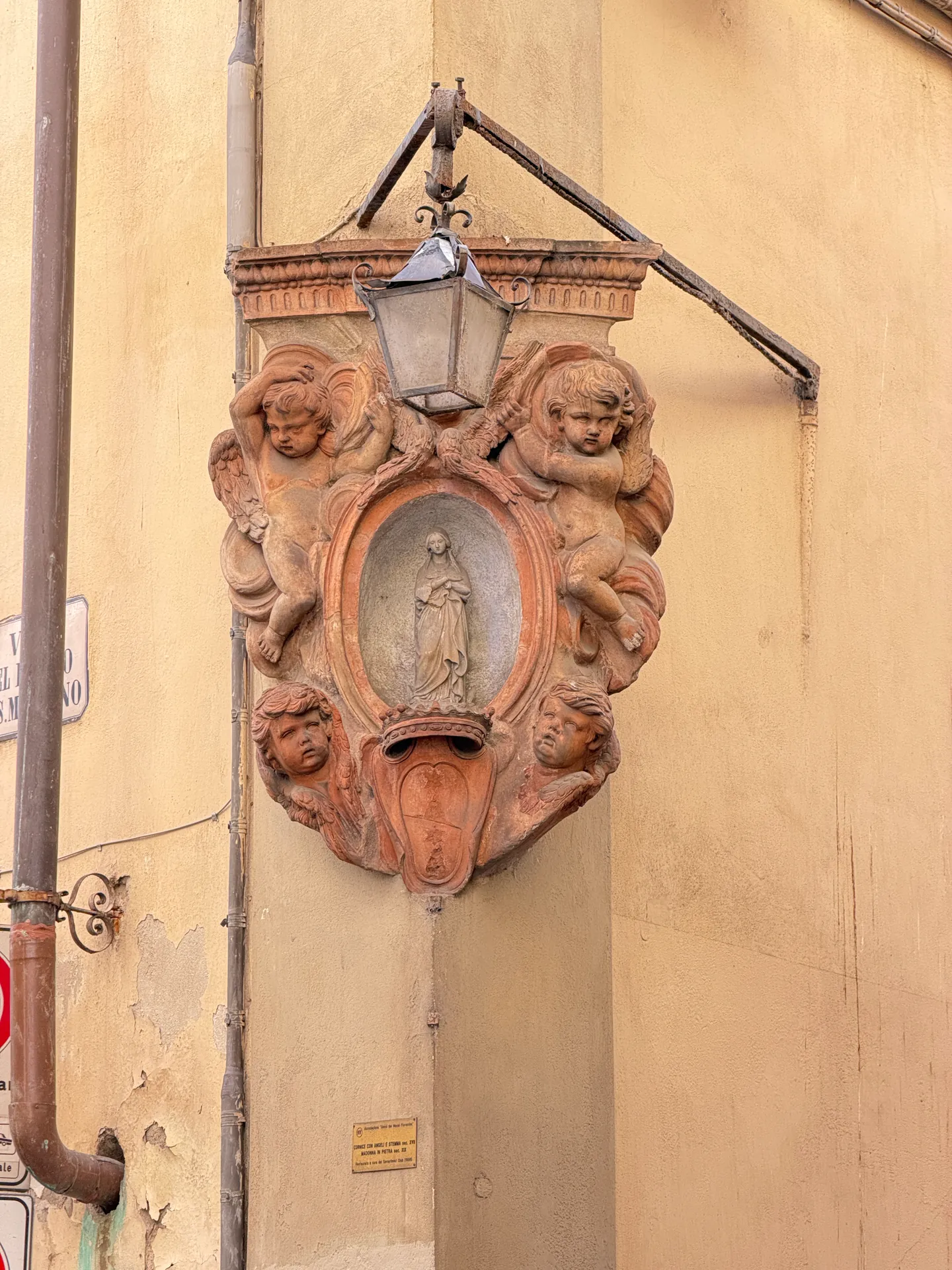 A terracotta street tabernacle on a corner, putti around a standing figure with a lantern above.