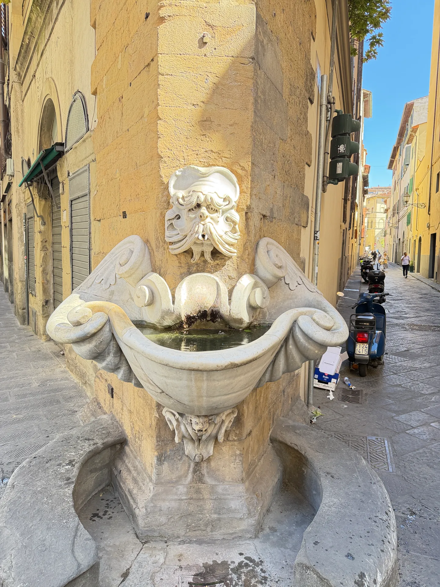A carved stone wall fountain with a mask and dolphins spilling into a basin, the street running past it.