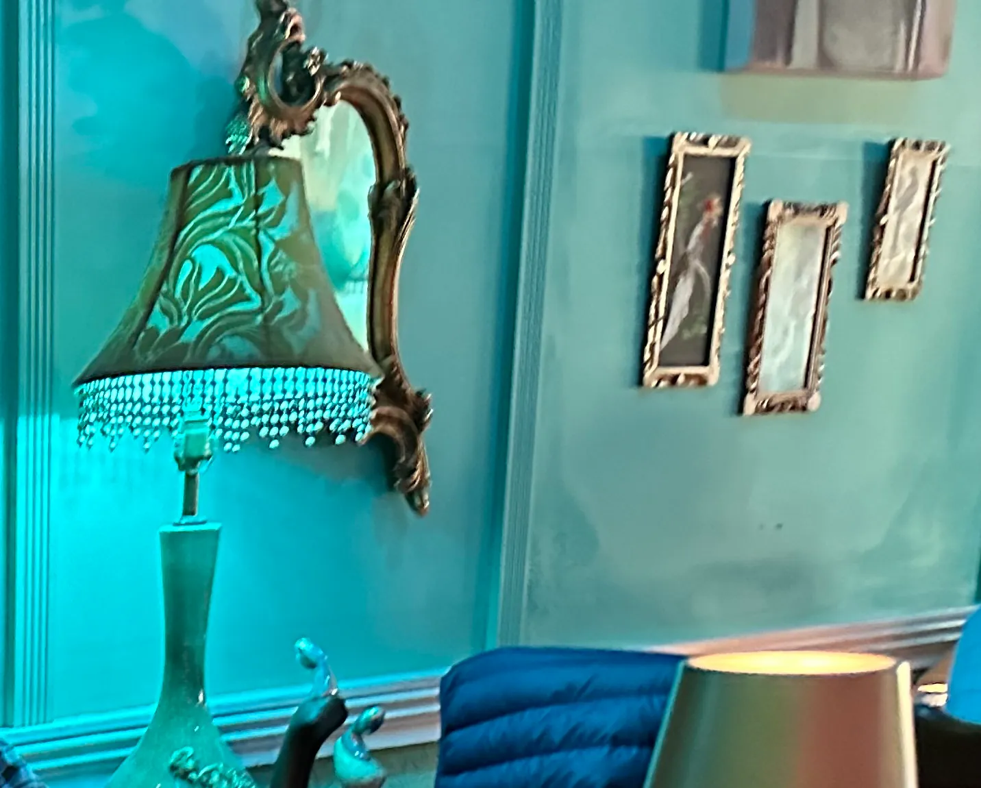 A fringed vintage lamp and gilt-framed pictures on a teal wall above a blue velvet banquette.