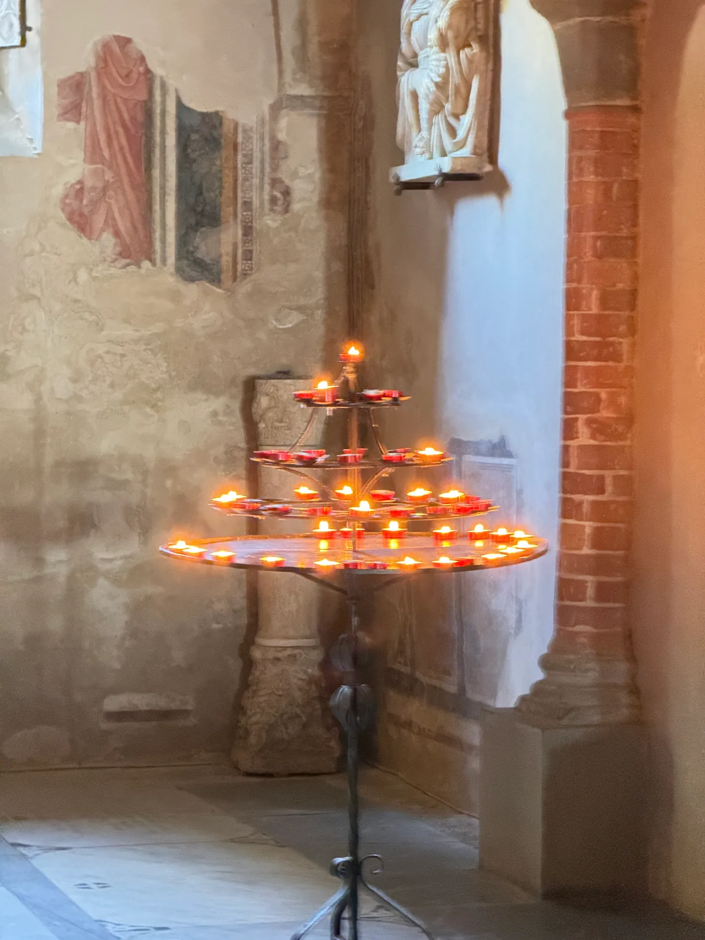 A votive candle stand burning in a corner chapel of San Miniato, a fresco fragment on the wall behind.