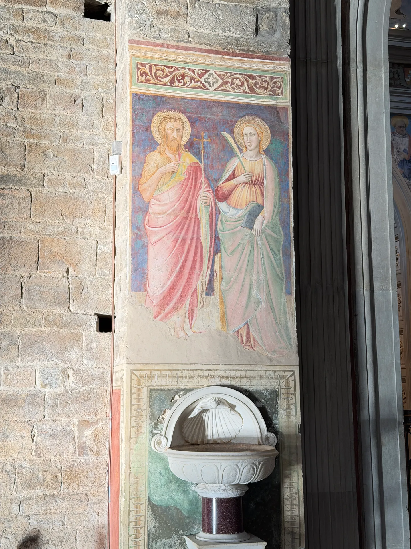 A stone holy water font against a nave pier of San Miniato, with a fresco of two saints on the wall above it.