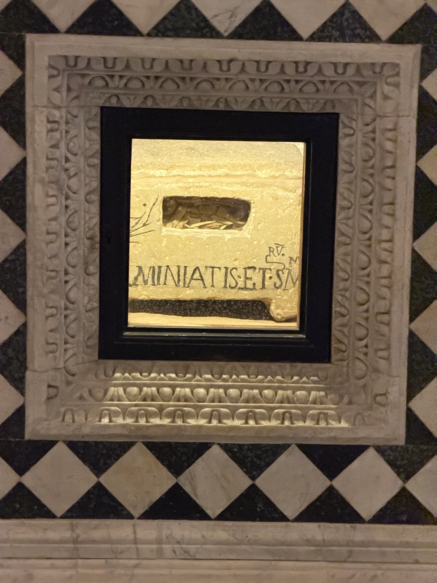 A gilded opening in the altar front of San Miniato's crypt, a carved slab behind it bearing the martyr's name.