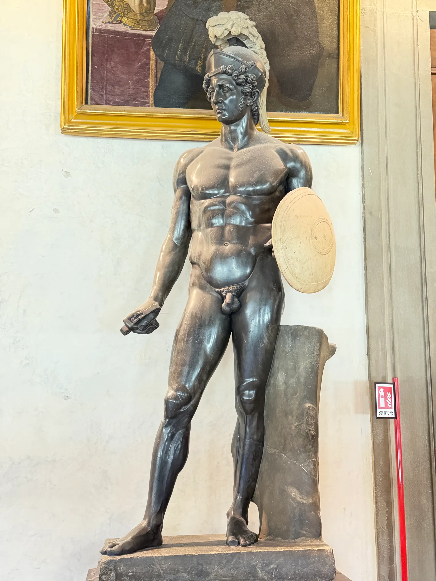 A dark bronze standing male nude on its plinth, a gilt framed painting on the wall behind.