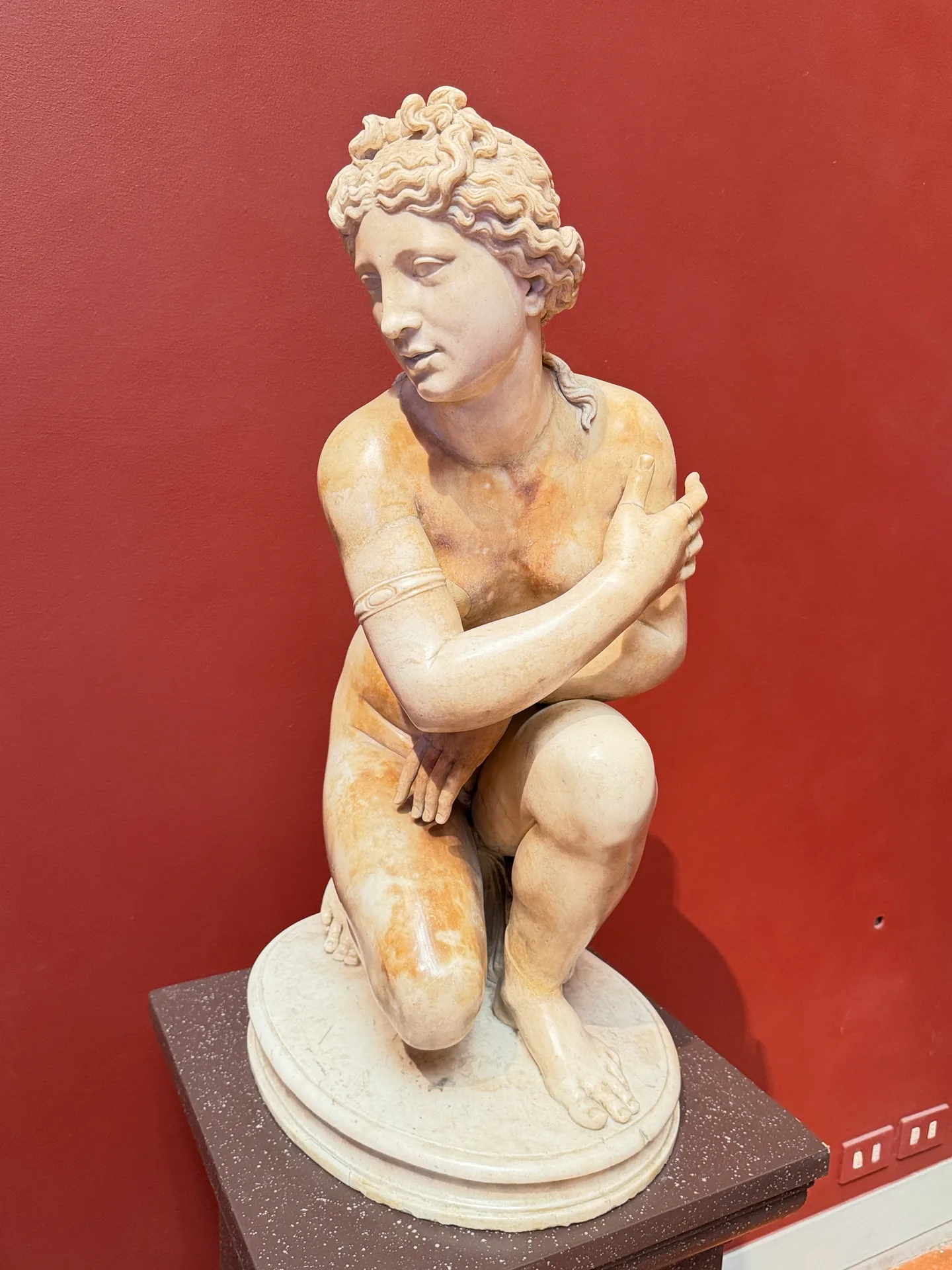 A Roman marble of a curly haired youth kneeling with his arms folded across his chest.