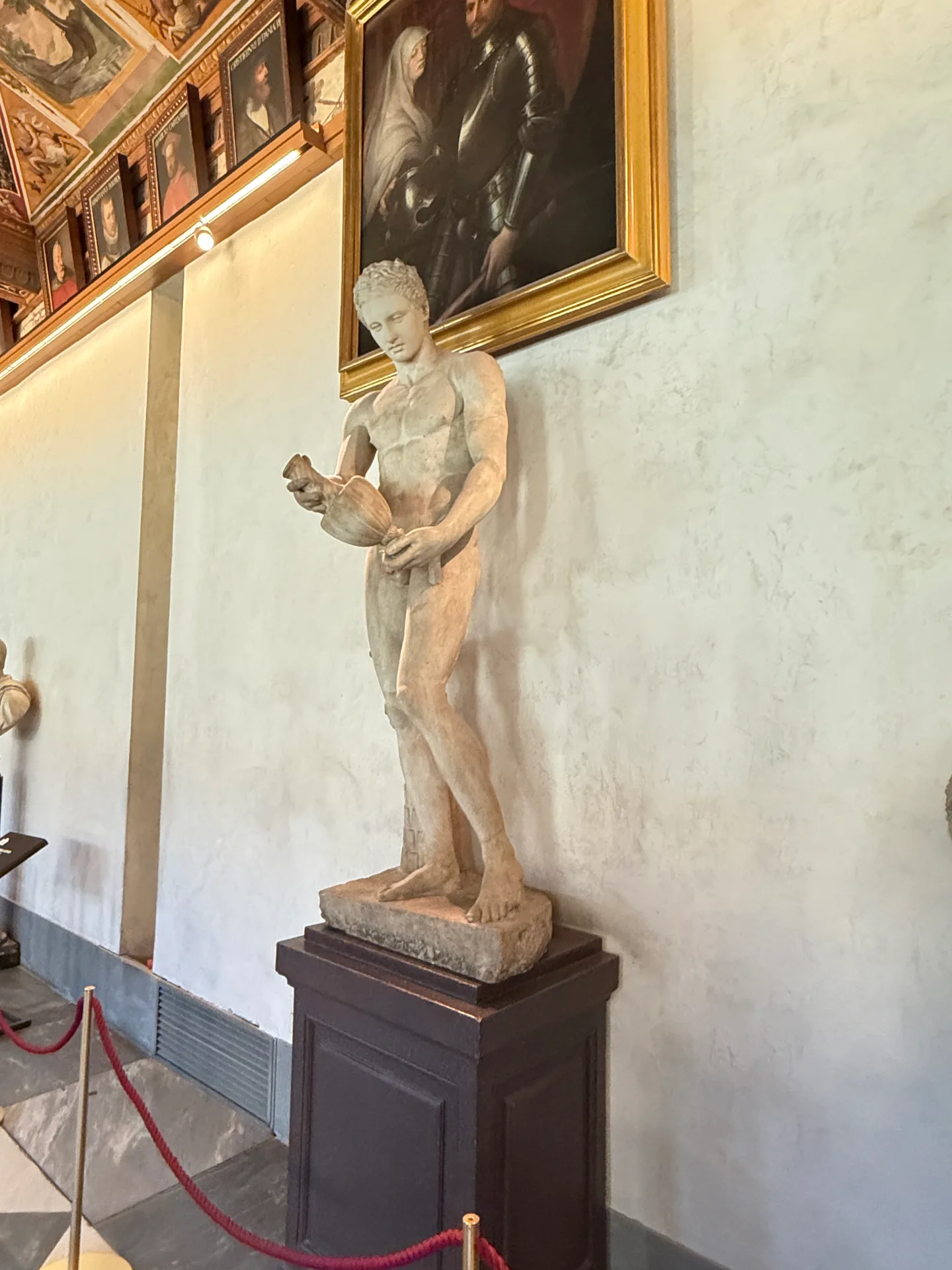 An antique marble of a standing nude youth with a tree trunk support, on a wooden plinth in the First Corridor.