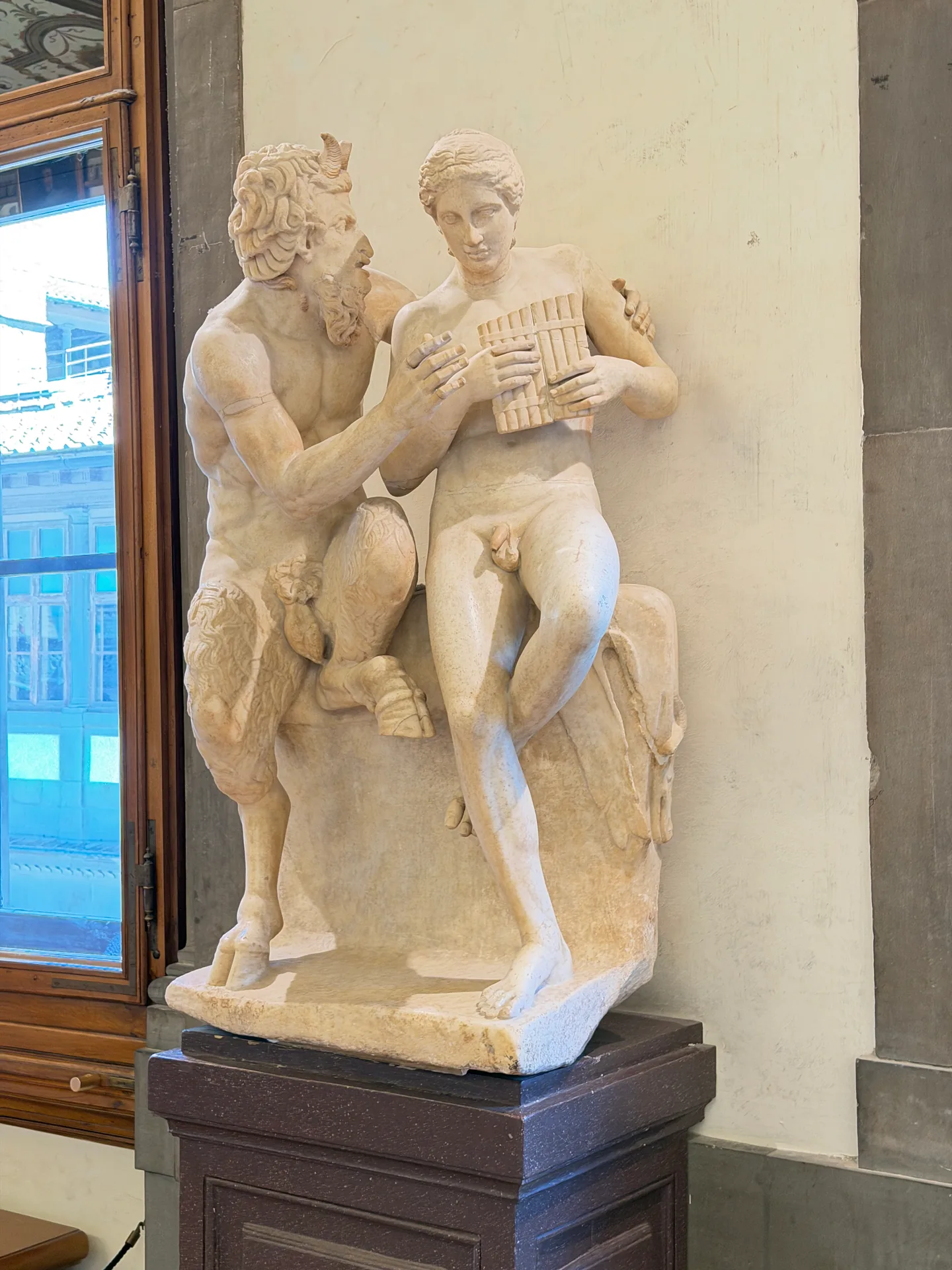 Two nude male figures carved as one group, standing on a dark plinth in the Uffizi corridor.