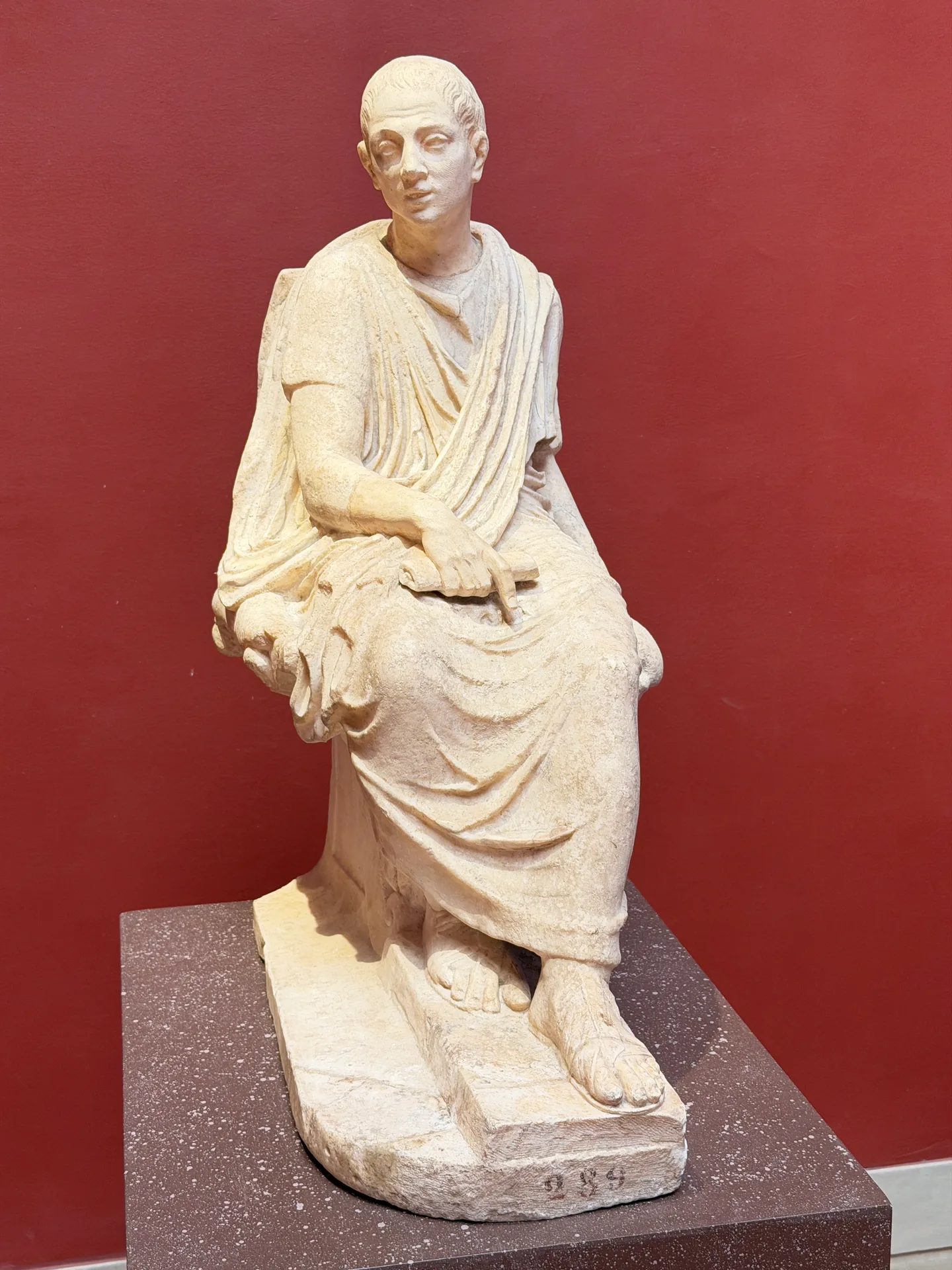A seated antique marble of a bearded man in chiton and himation, one hand on his knee, on a porphyry coloured plinth.