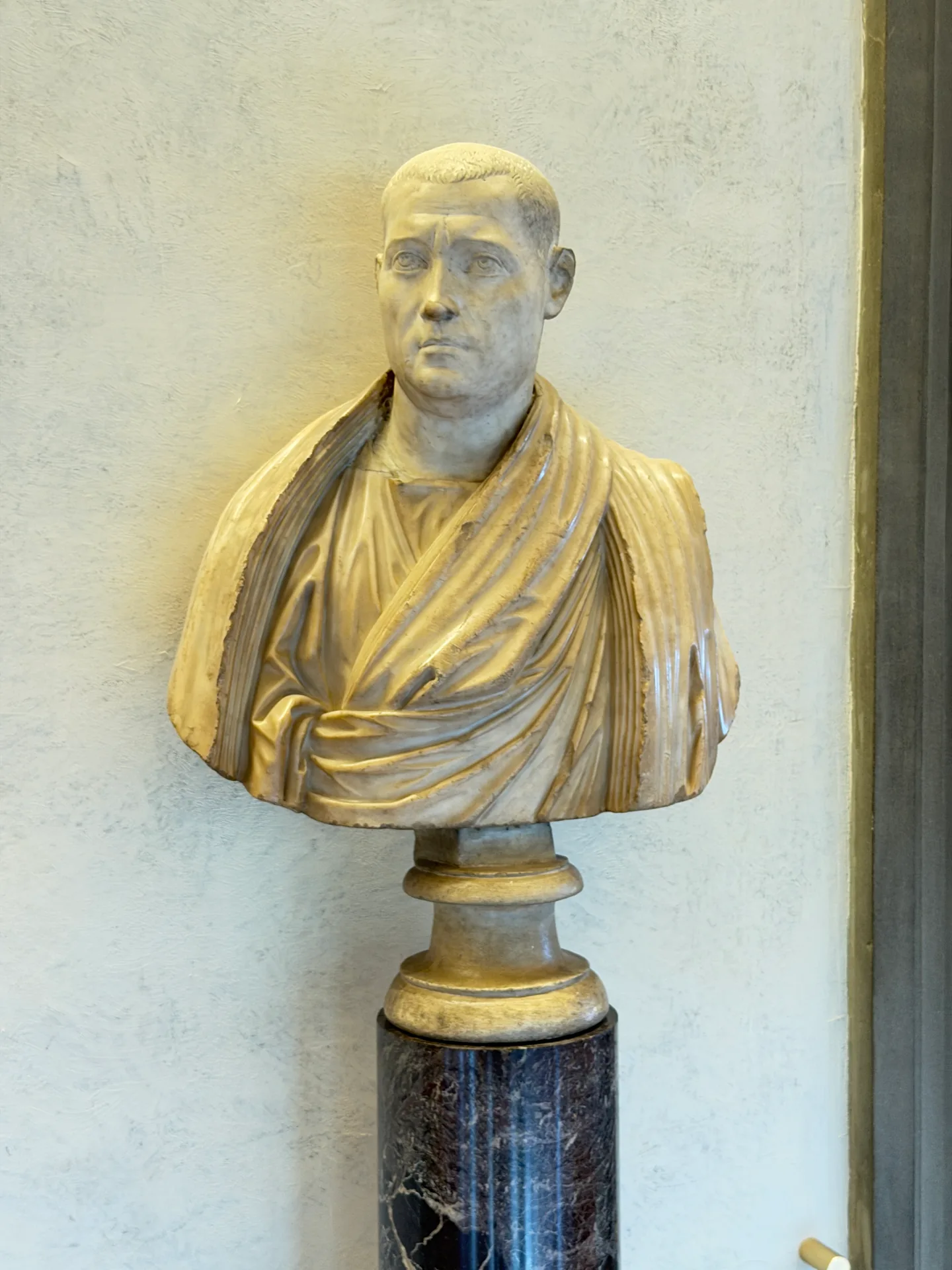 A Roman portrait bust in a toga, set on a dark marble column against a plain wall.
