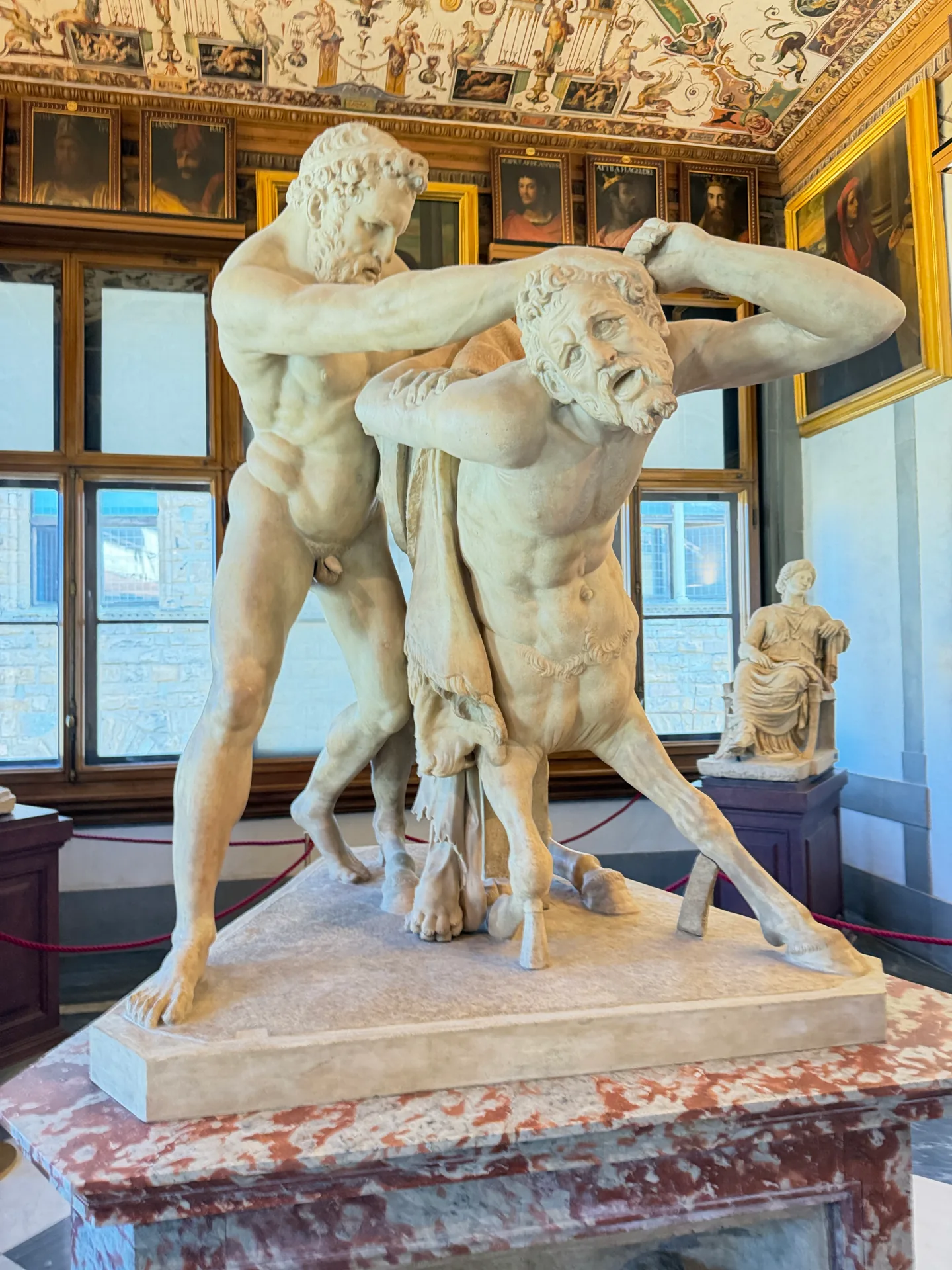 An antique marble group of two figures on a coloured marble base, lit from the corridor windows behind.
