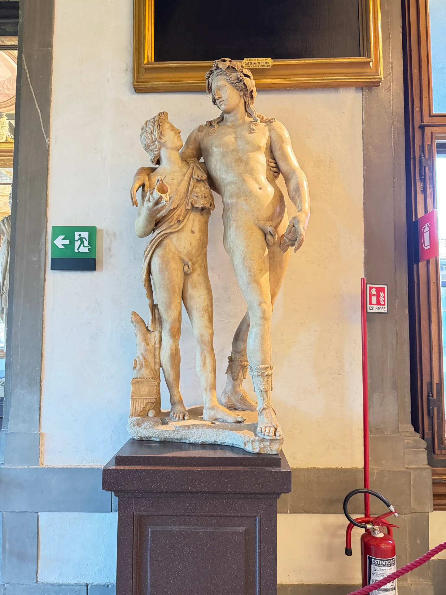 A marble group of a wreathed nude youth with a smaller draped companion, against a corridor wall.