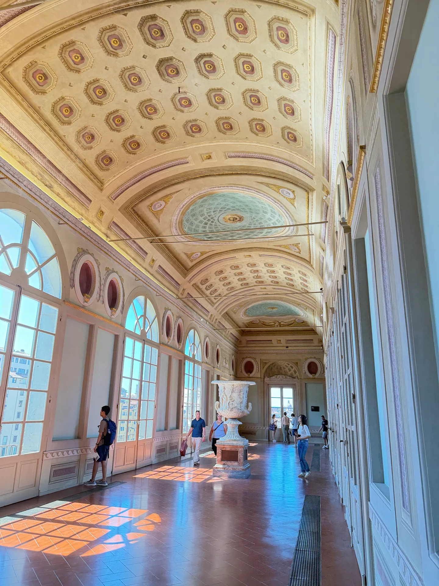 A first floor corridor of the Uffizi under plain barrel coffering, sunlight falling in patches on the wood floor.