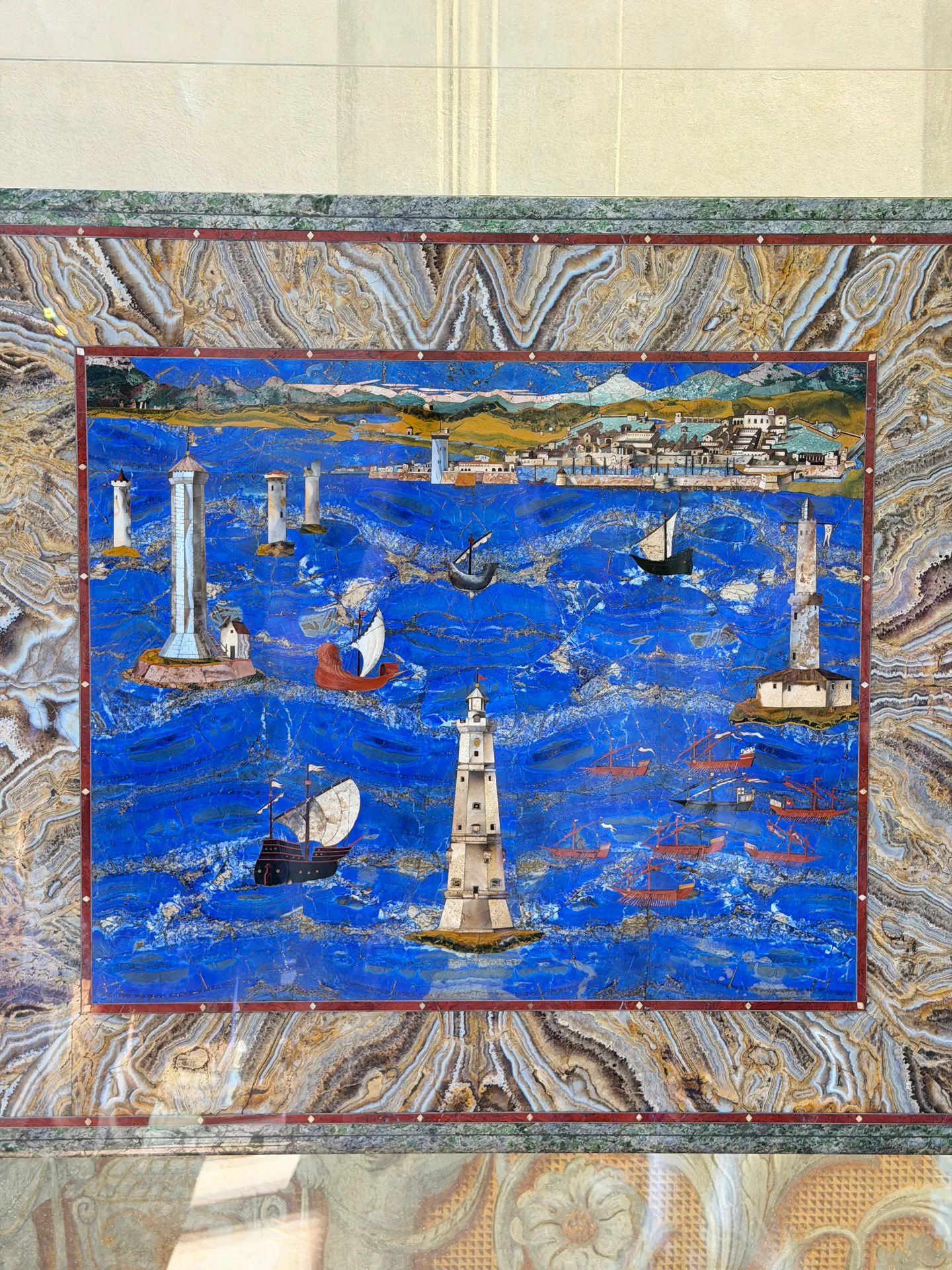 A pietre dure panel of a harbour, its lighthouse and shipping inlaid in coloured stone on a lapis ground.