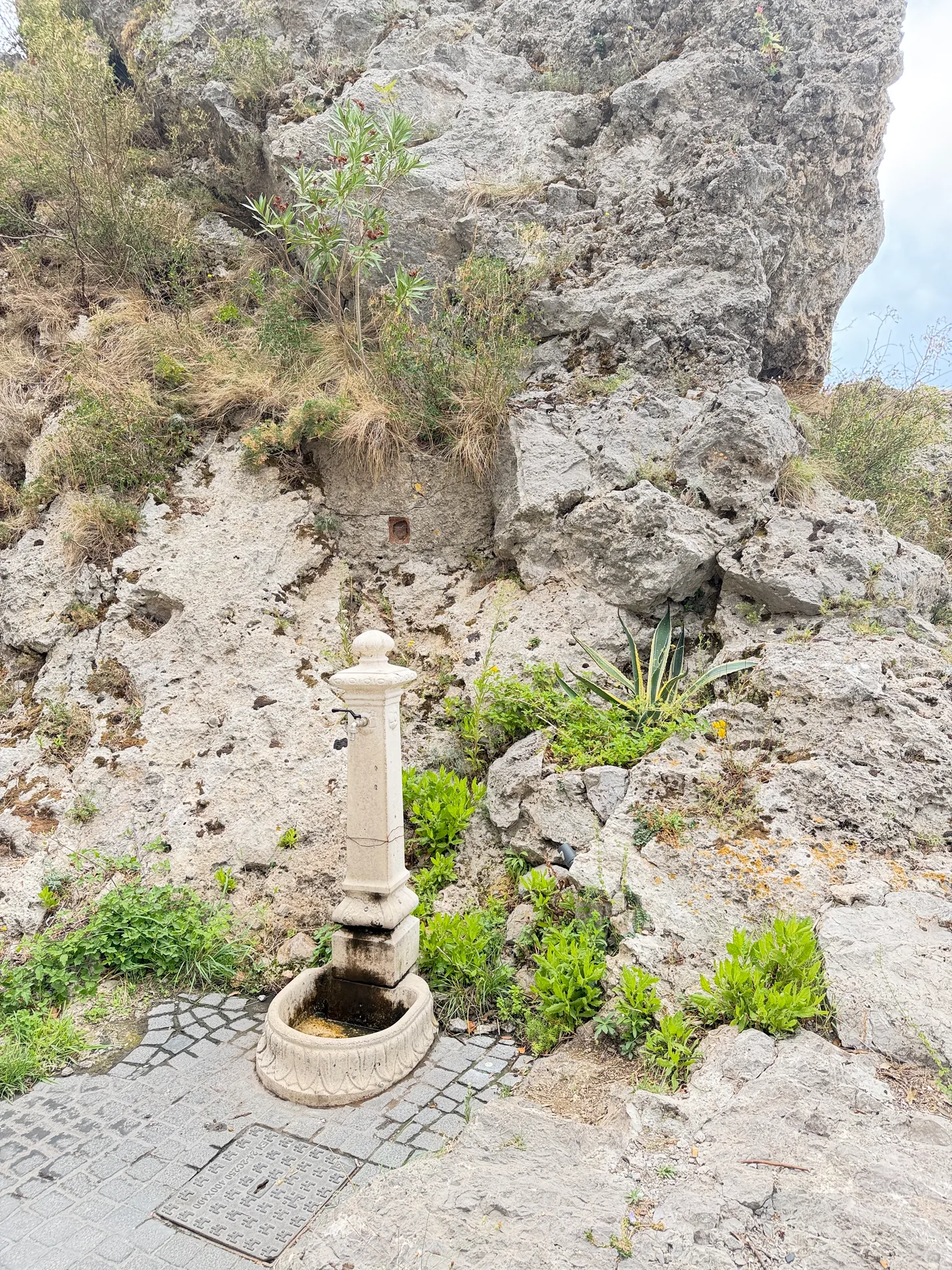A small stone fountain with a spout and basin built into a rock face beside a paved road