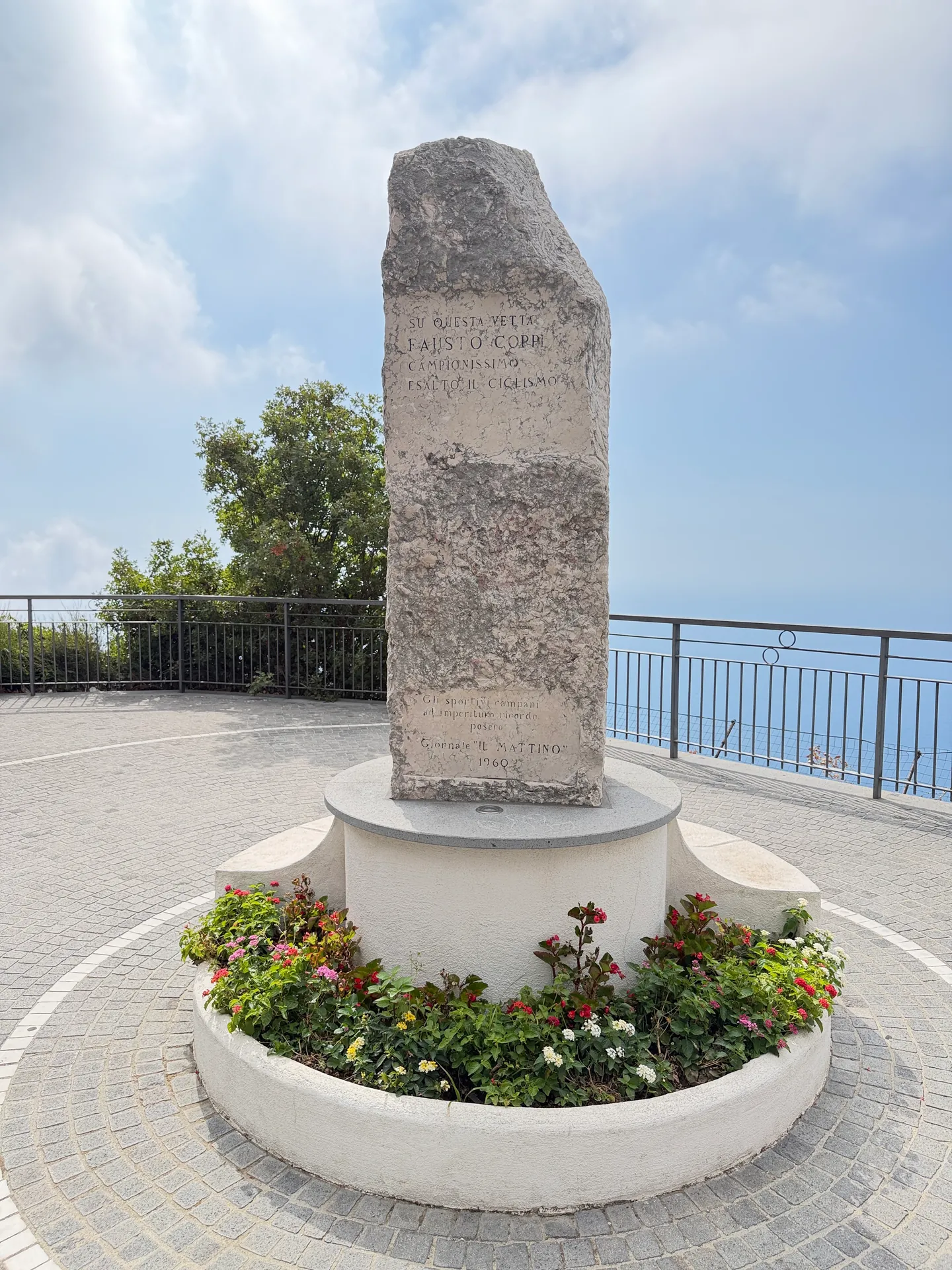 A tall stone stele on a paved belvedere in a round flowerbed, the sea visible beyond the railing