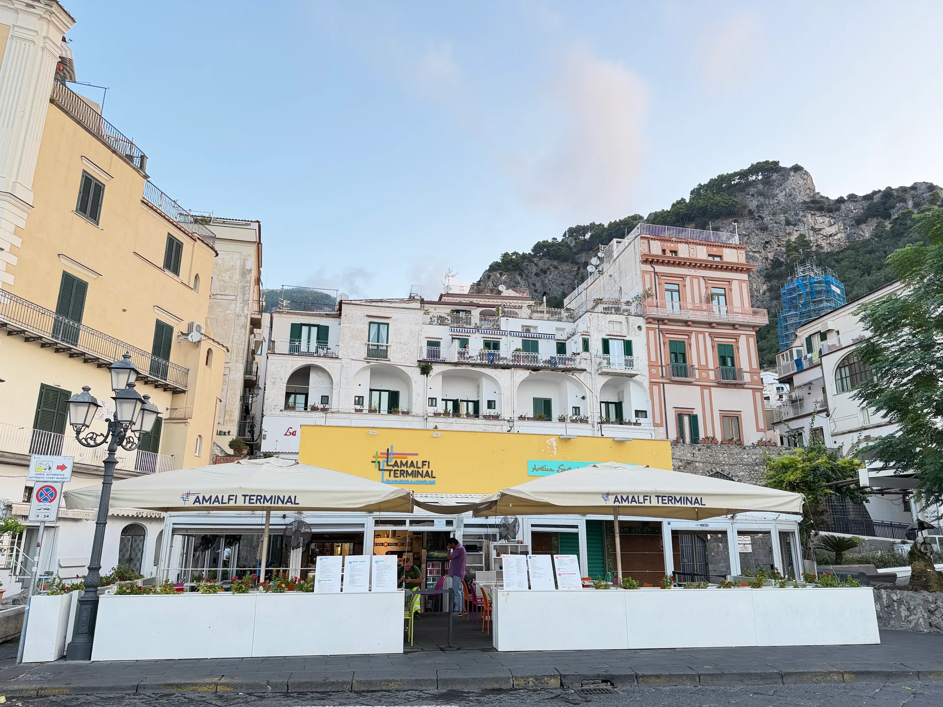The Amalfi Terminal on the seafront, cream umbrellas under a yellow sign, houses and hill behind