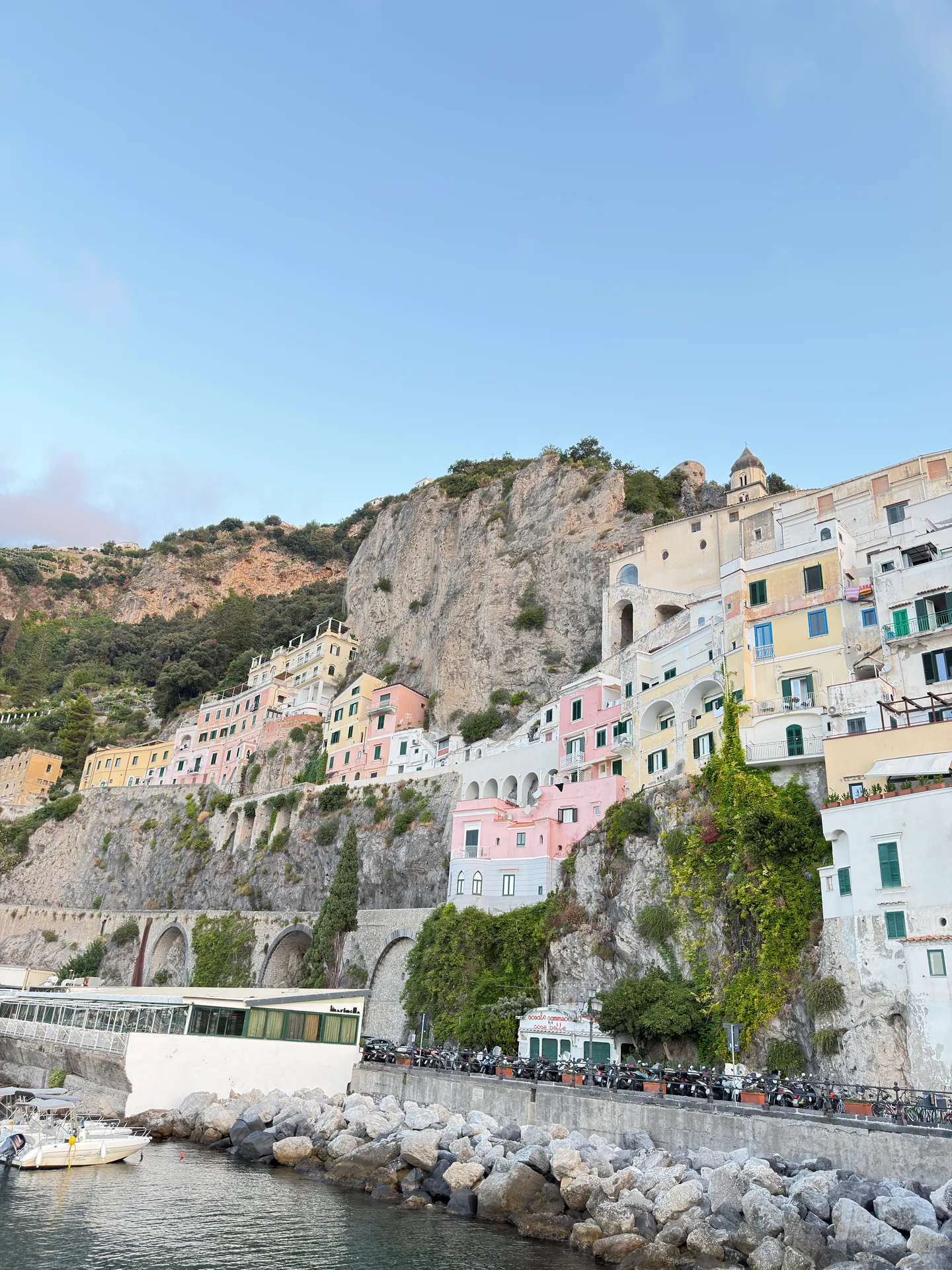 Pastel houses climbing a cliff above a road and a small boat on a rocky shore