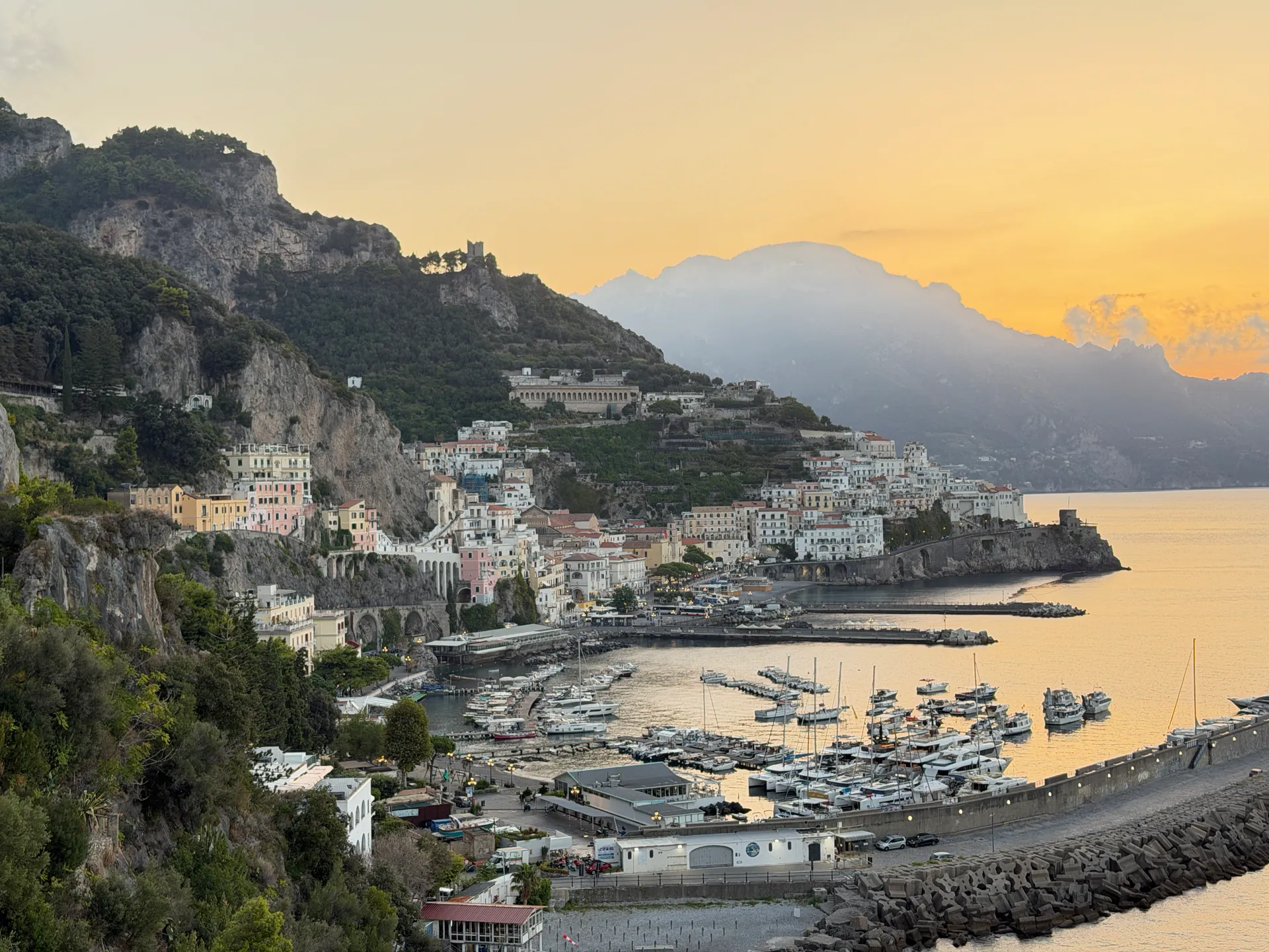 Amalfi harbour and its stone mole at dawn seen from above, the town on the hillside under a gold sky