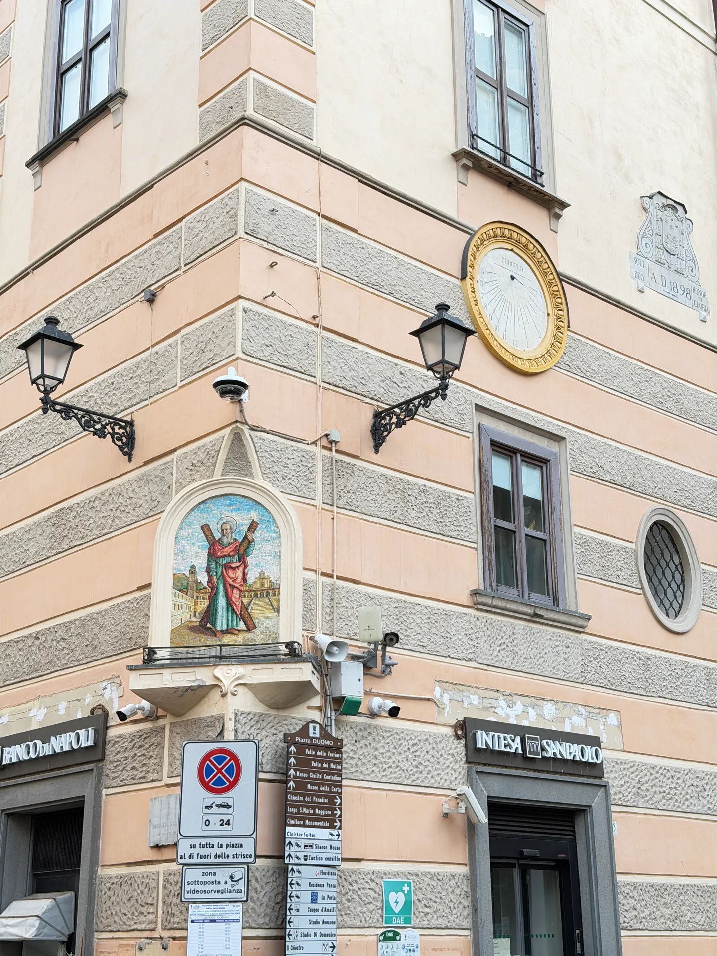 Building corner on Piazza Duomo with a mosaic saint in a niche, a sundial, a street plate and tourist signposts
