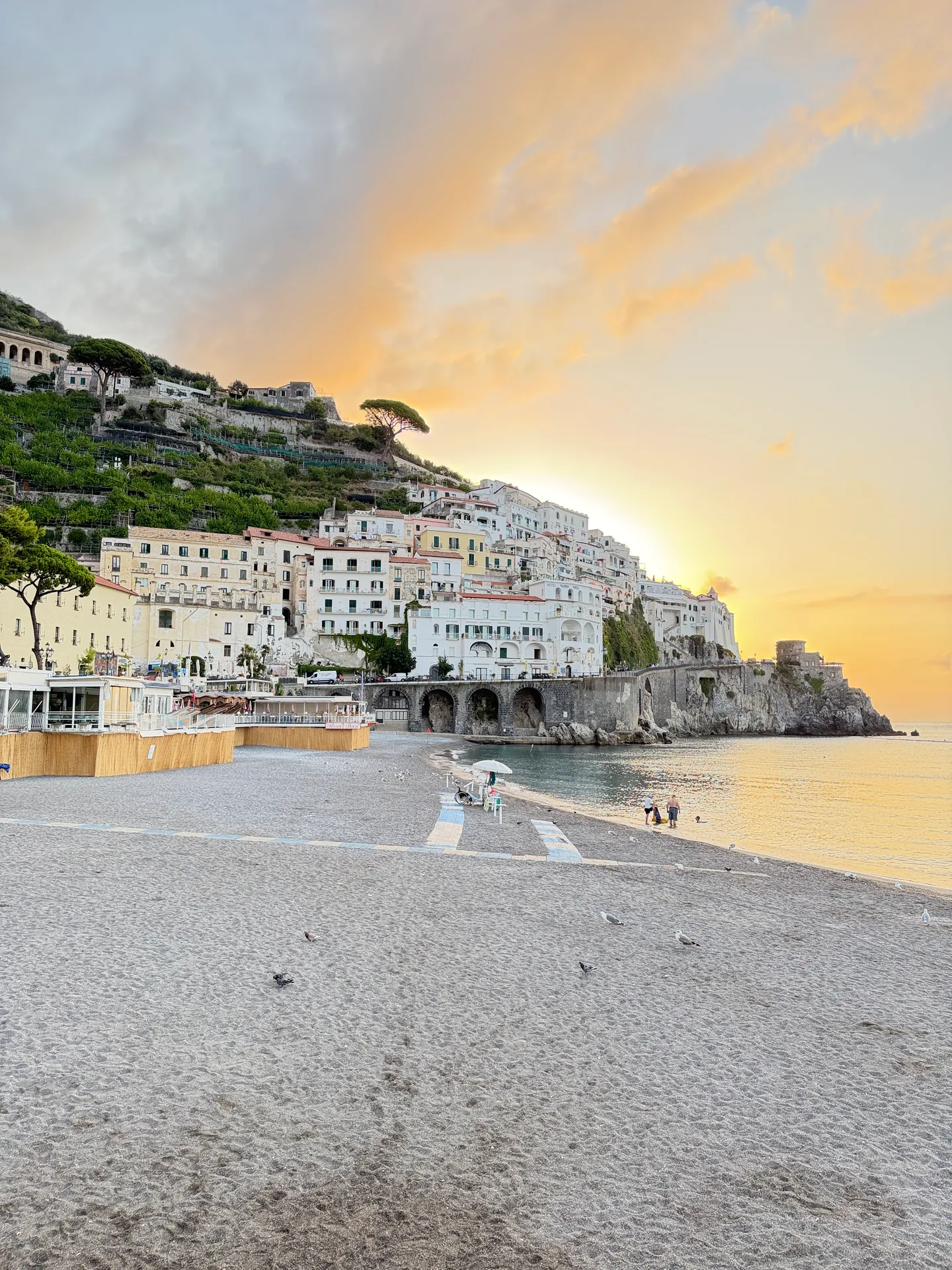 Grey sand beach at sunrise looking toward a headland with houses and a tower on it
