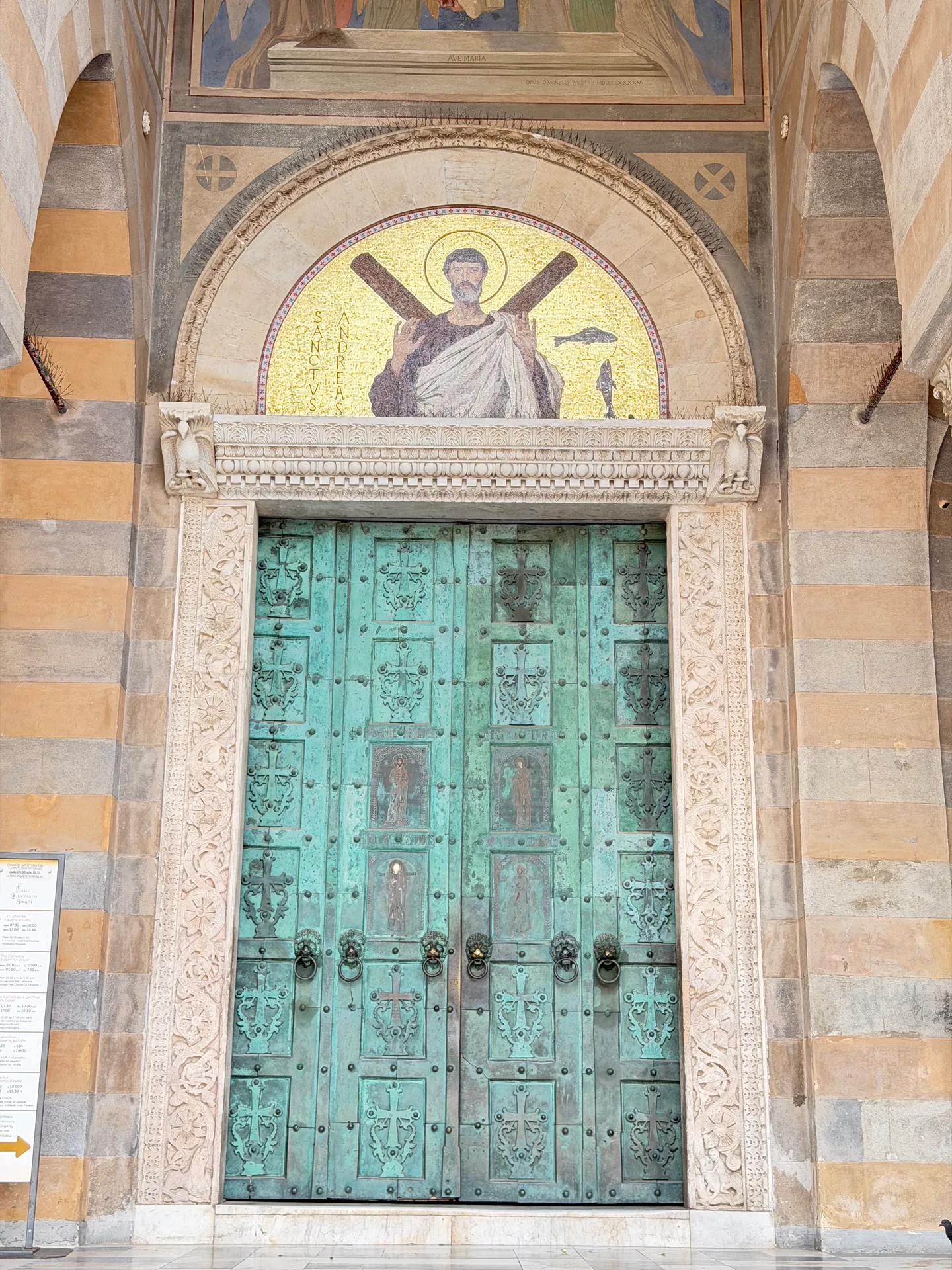 Green bronze door of Amalfi cathedral beneath a gold mosaic lunette, a notice board on the pier at left