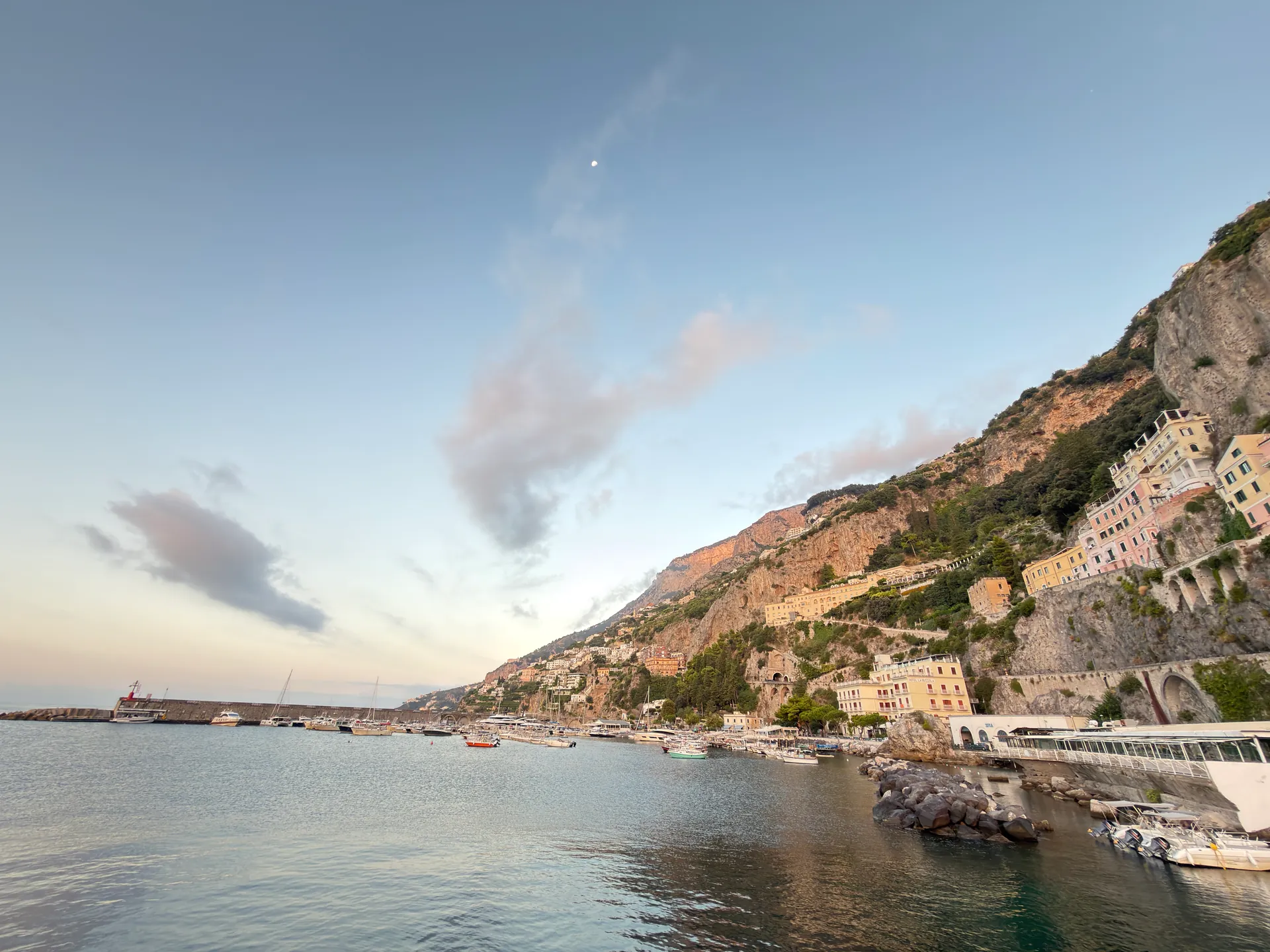 Calm morning sea in a cove with a stone harbour mole and a hillside town beyond