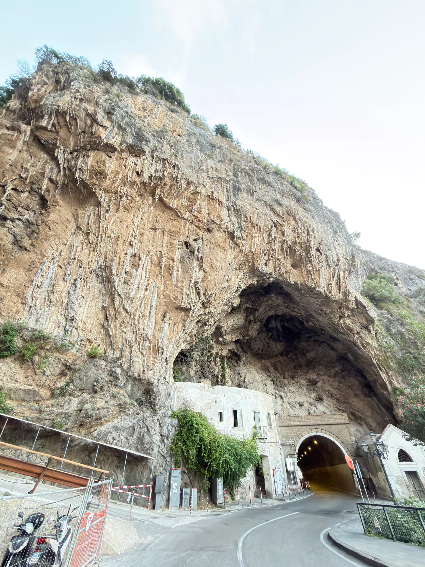 Road tunnel entrance beneath a tall limestone overhang, with a white building at its mouth and a road curving in