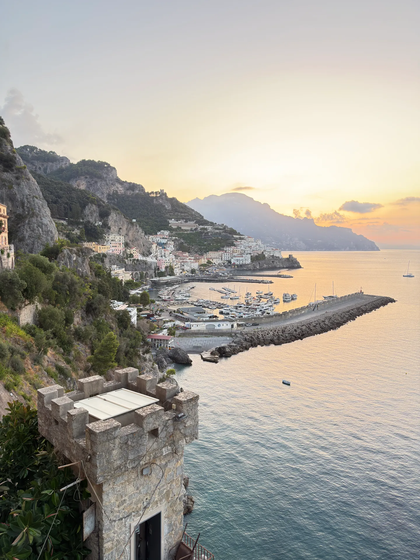 Crenellated stone tower above Amalfi harbour at sunrise, the mole and moored boats below