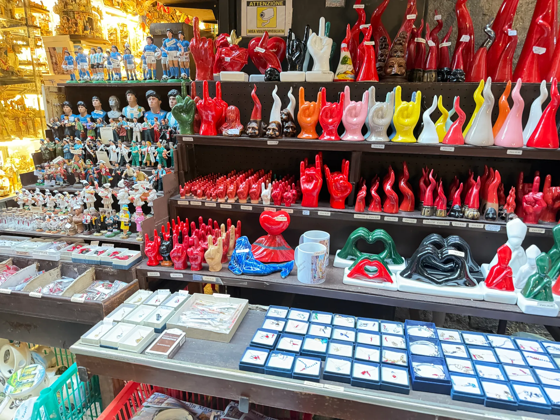 Rows of red horn charms and ceramic hands beside painted figures on San Gregorio Armeno