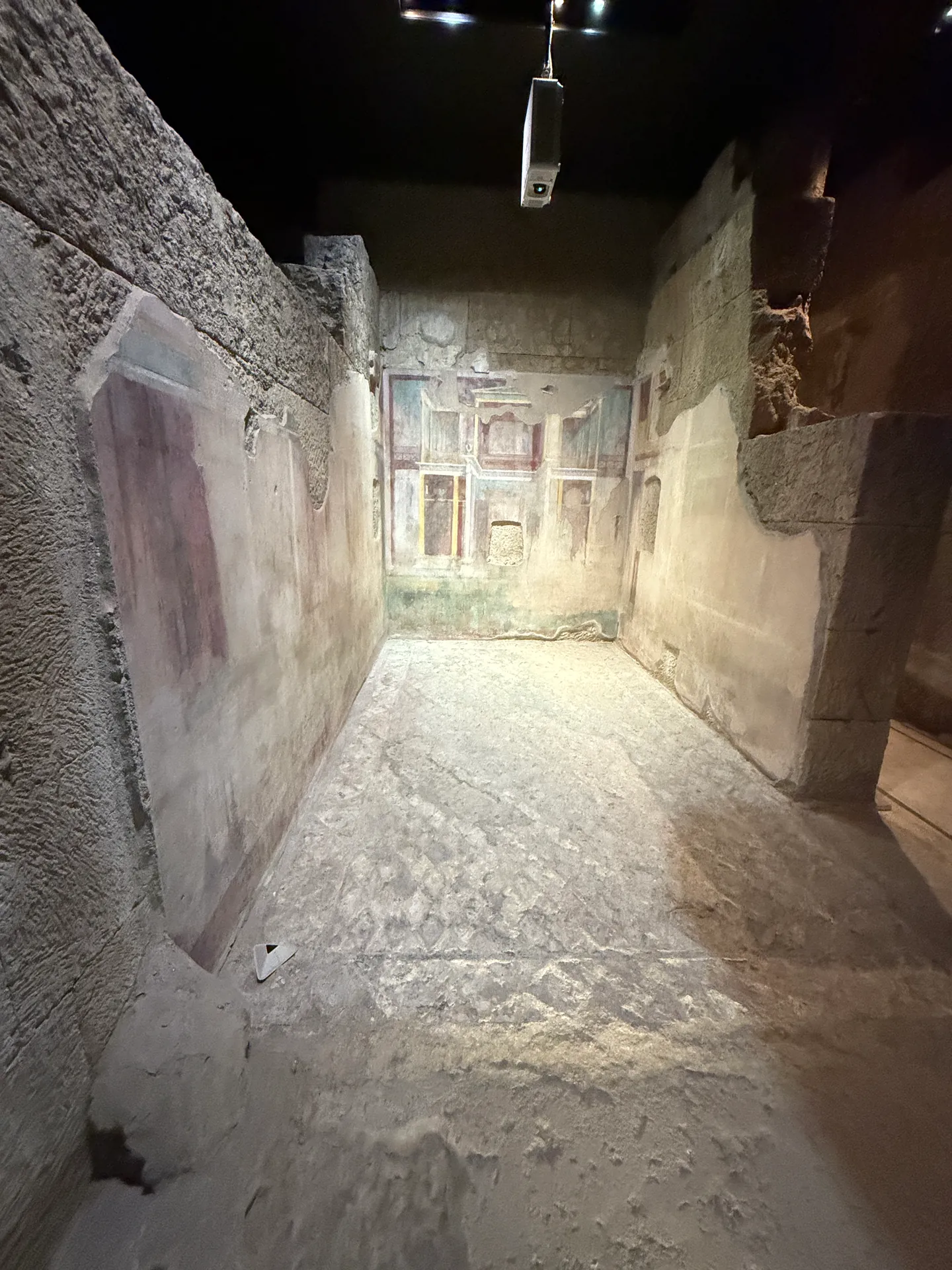 A room of the House of Augustus unlit, its far painted wall visible above a bare earth floor.